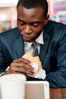 Afro-Amerikan iş adamı takım elbiseli, restoranda oturuyor, hamburger yiyor ve tablet kullanıyor.