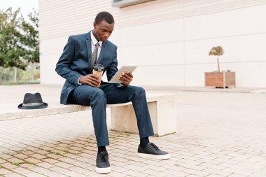 Genç ve zeki bir işadamı dijital tablet kullanıyor ve şehirdeki bir bankta bilgileri analiz ediyor. Dışarıda çalışmak ve mobil ofis işi kavramı.