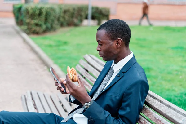 Hombre Afroamericano Comiendo Hamburguesa Parque Aire Libre Mientras Usa Teléfono — Foto de Stock