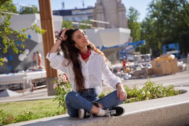 Genç, güzel, mutlu bir kızın sokak tarzı portresi. Yaz şehrinde oturuyor, uzun koyu renkli saçlara dokunuyor.
