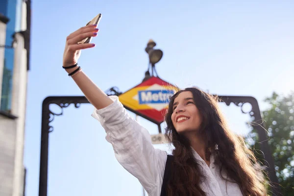 Madrid metro istasyonunun önünde gülümseyen genç bir kadının portresi. Kadın selfie çekiyor.
