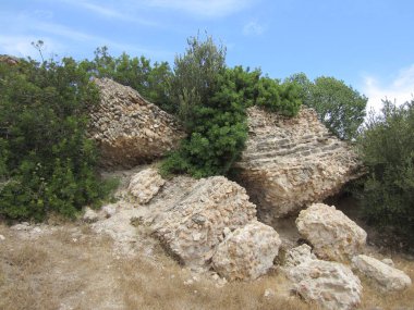 Büyük taşlar, yıkılmış bir binanın kalıntıları ve parlak mavi gökyüzüne karşı alçak bitkilerden oluşan yeşil çalılar..