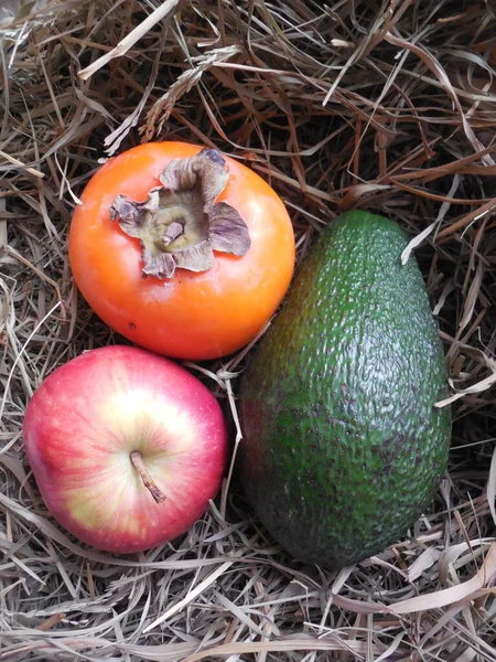 绿色鳄梨 橙黄色的柿子和成熟的多汁的红色苹果 背景美丽的干稻草 — 图库照片