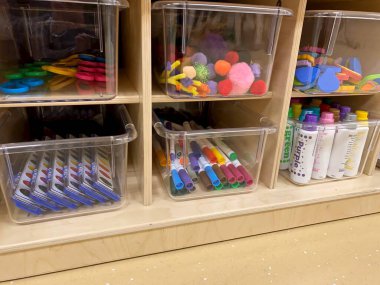 Early childhood education preschool classroom. Wooden shelves with art supplies in clear bins - scissors, paints, markers, pipe cleaners and shapes. No people. 