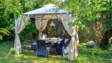 Bahçede, ağaçların arasında, taş bir duvarın karşısında tente bir çardak var. Güneş, yaprakların arasından doğar, masanın üzerinde yatar, çimenler, çatıda parlar. 