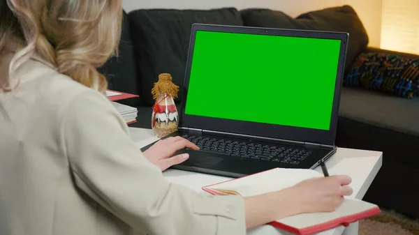 Chromakey Mockup. Boş yeşil ekranlı dizüstü bilgisayar kullanan ve yazan bir kadın. Ev işleri. Masada oturan dizüstü bilgisayardan notlar alıyor. Sarışın bayan.
