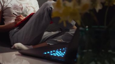 Dizüstü bilgisayarla not yazan genç bir kadın. Geceleri çevrimiçi ders alıyorum. Gece geç saatlere kadar oturma odasındaki kanepede oturup bilgi yazarak evde fazla mesai yapan serbest çalışan biri..