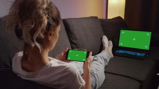 从肩膀上看过去 女人躺在沙发上 用手机和笔记本电脑 用绿色模拟屏幕按键观看内容 水平电话绿色屏风 使用智能手机回家 — 图库视频影像