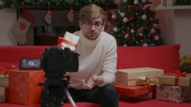 Beyaz kazak giyen yakışıklı bir adam Noel zamanı evde video blogu hazırlıyor ve online hediyeler alıyor. Teknoloji ve Tatil konsepti. Kış tatilinde hediye vermekle ilgili..