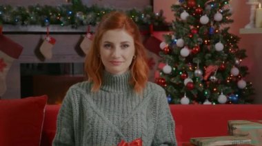 Gülümseyen güzel bir kadın portresi elinde Noel hediyesi kutusuyla arka planda Noel ışıkları süslemeli oda ve Noel ağacı. Kameraya bakan bir kadın, kızıl saçlı, mavi gözlü..