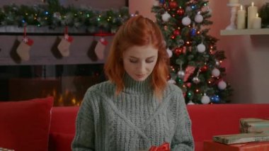 Gülümseyen güzel bir kadın portresi elinde Noel hediyesi kutusuyla arka planda Noel ışıkları süslemeli oda ve Noel ağacı. Kameraya bakan bir kadın, kızıl saçlı, mavi gözlü..