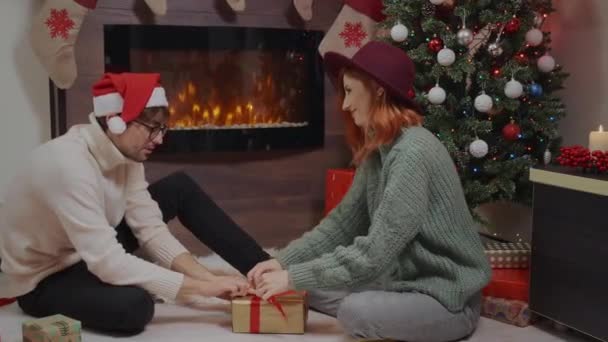 幸福的夫妻为家人和朋友包装圣诞礼品盒 在圣诞树下送礼寒假前夕令人惊奇 漂亮的女人 戴着帽子的红头发 英俊的男人 恋爱中的夫妻 — 图库视频影像