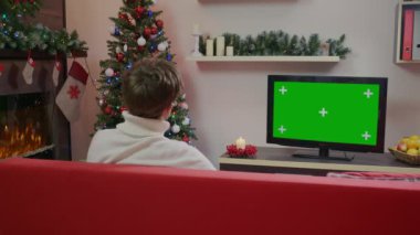 Süveter giyen adam oturma odasında kanepede otururken Yeşil Krom Ekran TV izliyor. Kış tatili dinleniyor. Noel tatili kavramı.