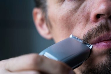 Bir adam sakalını ve bıyığını elektrikli jiletle keser. Tıraş olma zamanı, sakal ve bıyık bakımı, elektrikli traş makinesi.