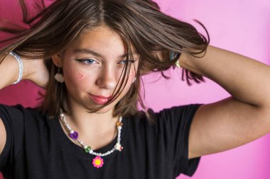 Saçlarını sallayan beyaz güzel bir kızın portresi, siyah bir tişörtle pembe bir arka planda poz vermek, yanaklarına kalpler çizmek.