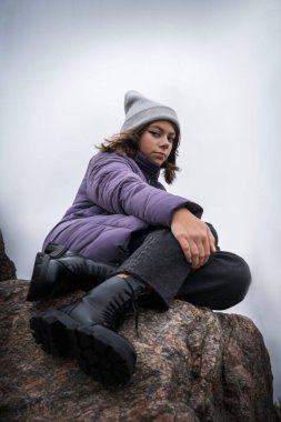Siyah saçlı, modern, kalın örgü şapkalı, mor önlüklü, siyah kot pantolonlu ve çıplak bir taşın üzerinde oturan siyah kabarık deri ayakkabılı hoş, modaya uygun beyaz bir kızın portresi.