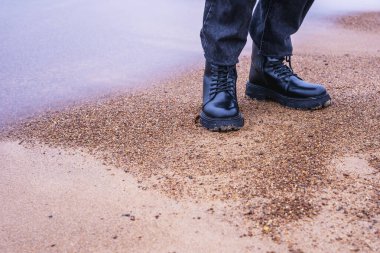 Siyah deri çizmeli ayaklar ve denizin kıyısındaki kumsalda duran siyah modaya uygun kot pantolonlar.