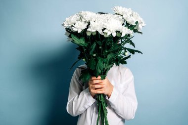 An unrecognizable boy in a white shirt holds a bouquet of white flowers in his hands, covering his face with flowers