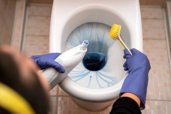 Women's hands cleaning a toilet bowl in a bathroom. A woman cleans the toilet in the bathroom using detergent, a brush and rubber gloves. Cleaning and cleanliness concept.