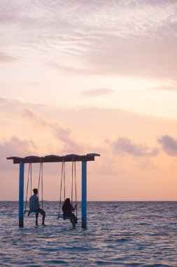 Maldivler 'in sığ denizinde salıncakta sallanan ve gün batımında güzel pembe gökyüzüne bakan iki çiftin resmi.