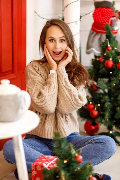 身穿米色针织保暖毛衣和蓝色牛仔裤的年轻女子在圣诞装饰品的背景下欢快地打开礼物 — 图库照片