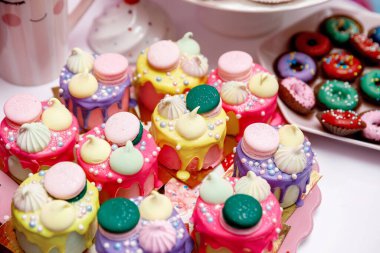 Masanın üzerinde hamur boncukları, krema ve kurabiyelerle süslenmiş renkli çörekler ve pastalar var.