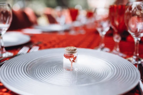 透明な小さな瓶の中の小さな赤い花が赤いテーブルクロスとテーブルセッティングのアイテムを背景に銀板の上に立っています — ストック写真