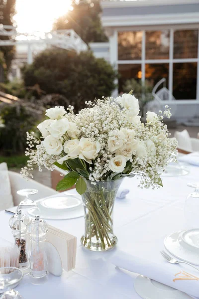 白色桌布上的桌子上有一束白玫瑰和吉普赛葡萄酒 背景上有餐具 — 图库照片