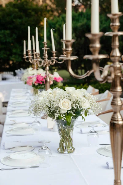 白玫瑰和吉普赛葡萄酒的花束 放在白桌布上的大烛台上 以花园的绿叶为背景 装饰有餐具 — 图库照片