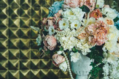 Yapay güllerin, şakayıkların ve ortancaların suni incilerle süslenmiş çiçek düzenlemesi duvardaki altın panellerin arka planına karşı