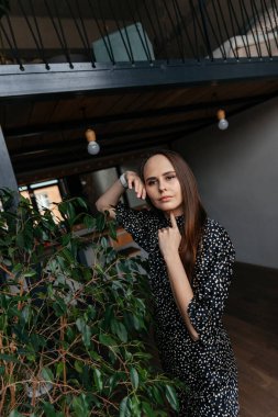 Puantiyeli bir elbise giymiş bir kadın yeşil bir çalılığın yanında duruyor oda dekorasyonunun arka planında.