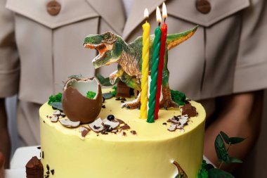 Bir adam elinde üç farklı renkte mum, bir çikolata yumurtası ve bir dinozor heykelciği ile süslenmiş sarı bir pasta tutuyor.