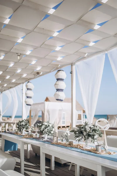 restaurant hall on the sea coast, decorated with paper lamps and white fabrics. tables are decorated with floral arrangements of white flowers and eucalyptus leaves, as well as candlesticks with white candles