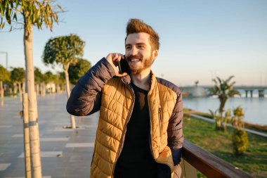 Genç adam telefonda konuşuyor, paltolu genç adam telefonda konuşuyor, güneşli bir kış gününde palto giyen bir adam ve soğuk hava.