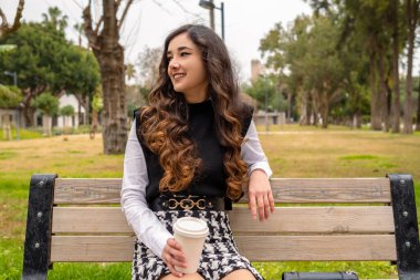 Geleceğe gülümseyen mutlu bir kadının portresi. Elinde bir fincan kağıt kahve tutan genç bir kadın. Parkta geleceğin hayalini kuran gülümseyen hoş bir genç kadın. Güzel kadın başka tarafa bakıyor, yeni zorluklar için heyecanlanıyor..