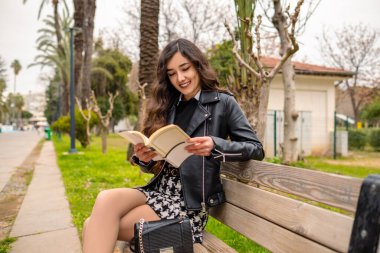 Kitap okumak, güzel bacaklı bir kadın parktaki bankta siyah beyaz elbise giyiyor. Bankta oturan ve parkta kitap okuyan genç ve mutlu bir kadın. Kişisel gelişim ve eğitim kavramı. Kadınlarda kendini geliştirme. Kızım parkta rahatla, bankta otur..