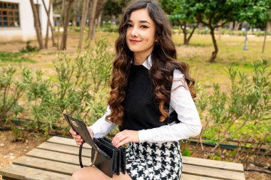 Siyah kazak ve beyaz gömlekle poz veren genç bir kadın. Çantada bir şey ararken kameraya bakan mutlu bir kadın. Dışarıdaki bankta oturuyor..