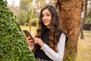 Poz veren, elinde telefonla kameraya poz veren genç bir kadın, yoğun çalışma saatlerine rağmen gülümseyen güzel bir yetişkin.