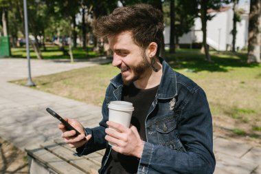 Gülümseyen, elinde kahve fincanı tutan genç adam bankta otururken telefonuna gülümsüyor ve eğleniyor.