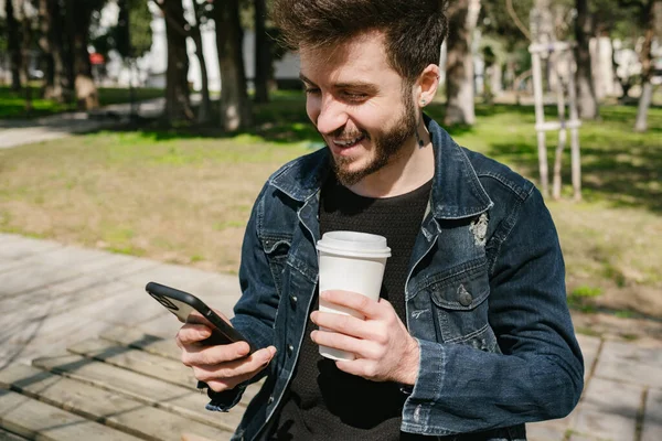Genç mutlu adam elinde bir fincan kahve tutarken telefon ekranına bakıyor. Dışarıda kahve içerken temiz hava soluyan bir adam. Rahatlamış ve sağlıklı bir yaşam tarzıyla mutlu..