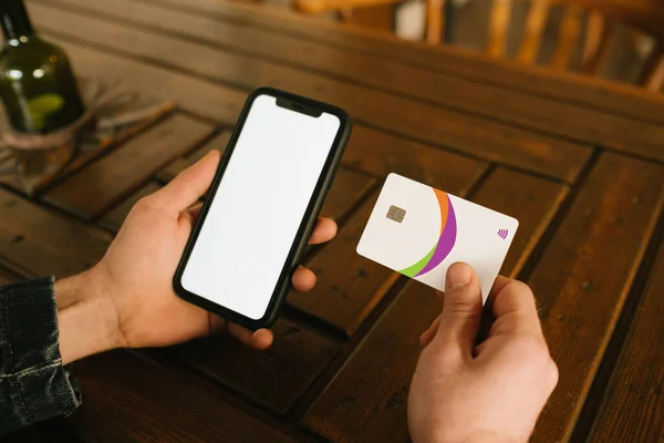 Bir kafede elinde kredi kartı ve telefonuyla oturmuş online alışveriş yapan genç bir adam. Çevrimiçi ödeme. Akıllı telefonu elinde tutan adam boş ekran ve kredi kartıyla işlem yapıyor.