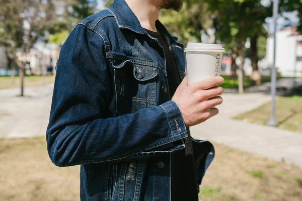 Denim ceket, kot ceketli kahve fincanı tutan genç adam, açık havada eğleniyor ve kahvenin tadını çıkarıyor.