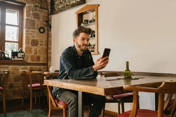 Genç adam kollarını ahşap bir masaya koyar ve elindeki telefona, eski bir kafeye, nostaljik bir bakışa, ahşap tonlara ve estetik bir yere bakar.