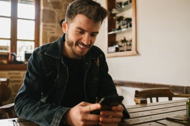 Kot elbiseli genç adam kahve masasında oturuyor ve kafede gülümseyerek telefonda mesajlaşıyor. Mutlu adam sosyal medya platformunda kayıyor ya da mesaj yazıyor ya da video izliyor.