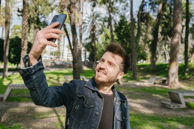 Şehir parkında dikilirken kendi portresini çeken yakışıklı bir genç. Gülümseyen adam selfie çekiyor..