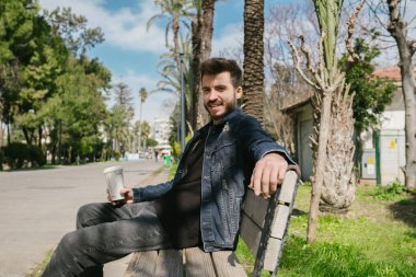 Kameraya poz veren, güzel havanın tadını çıkaran genç adam elinde koyu mavi gökyüzü olan bir kahve fincanı tutuyor.