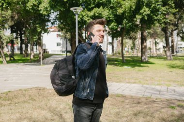 Genç adam seyahat ediyor, tam bir sırt çantasıyla yola çıkmak üzere, kot ceketli adam telefonda konuşuyor, ağzına kadar dolu bir sırt çantası.