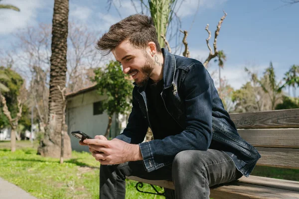 Açık havada cep telefonuyla oturan gülümseyen bir gencin yan portresi. Mutlu adam olumlu mesaj ya da video mesajı okuyor ya da çevrimiçi video izliyor. Açık havada mobil oyun oynanıyor.