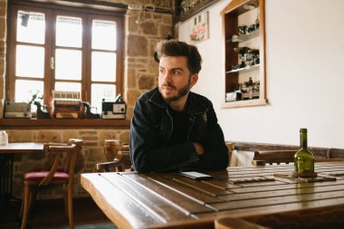 Düşünceli bir adam eski bir yerde oturmuş kahvesini bekliyor, kot ceketli sakallı adam kollarını kavuşturmuş ve yüzü sıkıldığını gösteriyor, telefon masanın üstünde.