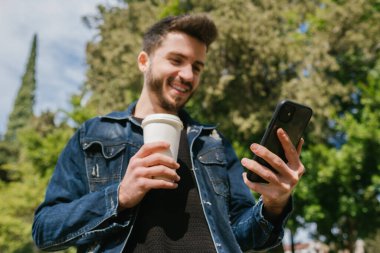 Açık havada elinde kağıt kahve fincanı tutarken cep telefonu ekranına bakan kot pantolon giyen mutlu genç adam. Sosyal medyada kaydırma, video izleme veya arkadaşlara mesaj gönderme.